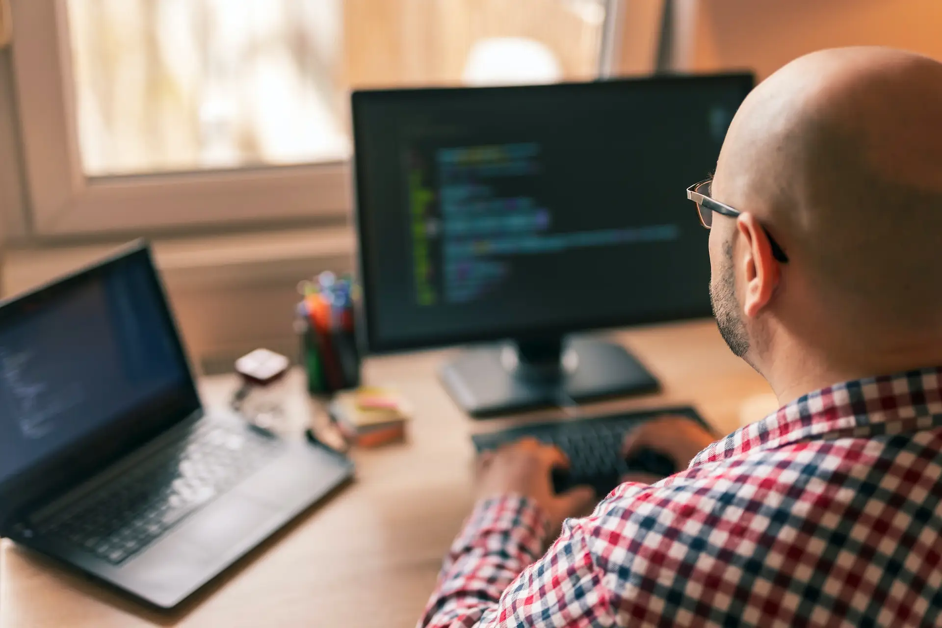 Person at a desk with a laptop and monitor displaying code, in a well-lit home or office setting.