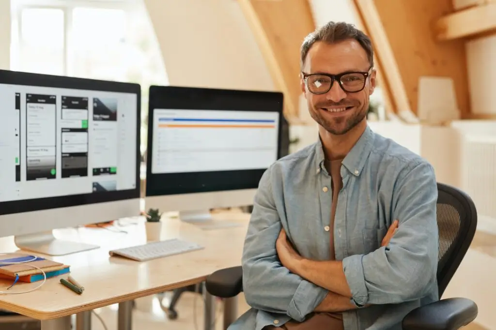 Person seated at desk with two monitors, wearing glasses and a blue shirt, smiling, well-lit office.